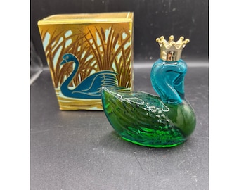 May include: A vintage glass swan perfume bottle with a gold crown stopper. The swan is teal and green, with a matching box featuring a swan illustration in teal and gold. The box has a gold background with a reed design.