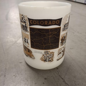 Puede incluir: Taza de cerámica blanca con un diseño marrón y negro que presenta un mapa de Colorado, la flor del estado y varios puntos de referencia. La taza dice "COLORADO" en letras negras.