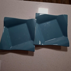 May include: Two teal square plates with a distinctive, folded design. The plates have a glossy finish and are placed side by side on a light-coloured surface. The plates are a modern addition to any dining set.