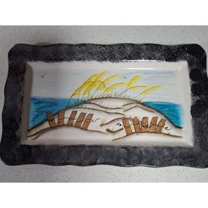 May include: Rectangular ceramic serving platter with a hand-painted beach scene. The design features a sand dune with tall yellow grass, a blue ocean, and a wooden fence. The border is black and grey.