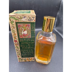 May include: A rectangular perfume bottle with amber liquid and a gold cap, standing next to its matching box. The box features a green background, a white swan illustration, and text referencing the Twelve Days of Christmas, marked 2 fl. oz.