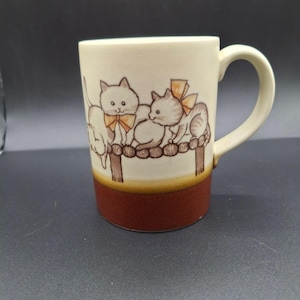 May include: Cream-colored ceramic mug with a brown base and handle. The mug features a charming illustration of three kittens with orange bows, perched on a wooden structure. The design is in a vintage style, perfect for cat lovers.