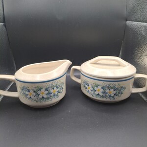 Op de afbeelding: Crèmekleurige melk- en suikerpotset met een blauwe streep en bloemmotief. De melkkan heeft een handvat en tuit, terwijl de suikerpot een deksel en twee handvatten heeft. Het bloemmotief heeft witte en blauwe bloemen.