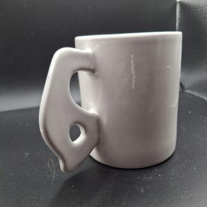 May include: A light gray ceramic mug with a unique handle design. The handle has a curved shape with a circular opening. The mug is cylindrical and appears to be made of a smooth, solid material.