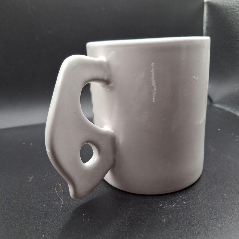 May include: A light gray ceramic mug with a unique handle design. The handle has a curved shape with a circular opening. The mug is cylindrical and appears to be made of a smooth, solid material.