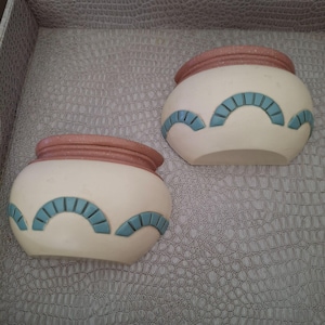 Op de afbeelding: Twee crèmewitte keramische potten met een ronde vorm. Elke pot heeft een roodbruine rand en een turquoise boogvorm rond de basis. De potten worden gepresenteerd op een getextureerd, grijs oppervlak.