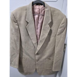 Könnte beinhalten: Ein beigefarbener Blazer mit Zwei-Knopf-Verschluss, fallendem Revers und zwei Vordertaschen. Der Blazer hat ein hellrosa Futter und hängt an einem schwarzen Kleiderbügel. Der Stoff hat ein dezentes Muster.