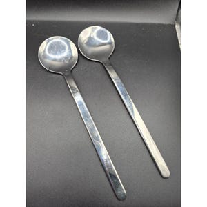 Könnte beinhalten: Zwei Edelstahl-Löffel mit runden Schalen und langen, schlanken Griffen. Die Löffel sind diagonal vor einem dunklen Hintergrund positioniert und reflektieren das Licht. Diese Utensilien eignen sich zum Servieren oder Essen.