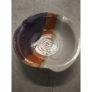 May include: Handmade ceramic bowl with a unique design. The bowl features a color-blocked pattern with sections of deep purple, orange, and off-white. A spiral design is in the center. The bowl has a small spout on the side.