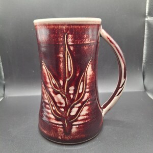 Può includere: Una tazza alta in ceramica bordeaux con un motivo a foglie. La tazza presenta un bordo e un manico bianchi, con un motivo a foglie strutturato in una tonalità più chiara di bordeaux. Il manico è curvo e attaccato al lato della tazza.