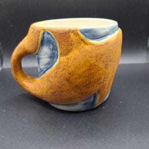 Może przedstawiać: Ręcznie wykonany ceramiczny kubek o unikalnym designie. Kubek ma teksturowaną, nakrapianą brązową powierzchnię zewnętrzną z niebieskimi akcentami. Wnętrze jest kremowe, a uchwyt jest zintegrowany. Odpowiedni do gorących lub zimnych napojów.