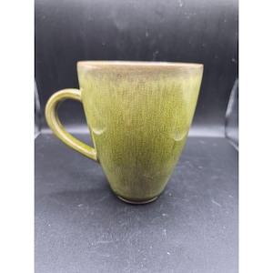 Könnte beinhalten: Eine grüne Keramiktasse mit gebogenem Henkel. Die Tasse hat eine strukturierte Oberfläche und einen dunkleren braunen Rand. Die Tasse ist leer und steht auf einer dunklen Oberfläche.