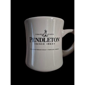 Puede incluir: Una taza de cerámica color crema con asa, que presenta texto negro y un gráfico de tipi. El texto dice: TRADE MARK PENDLETON SINCE 1863 PENDLETON WOOLEN MILLS + PORTLAND, OREGON. La taza tiene un acabado suave y brillante y una forma curva.