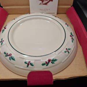 Puede incluir: Un plato de cerámica blanco con un diseño de bayas de acebo verdes y rojas alrededor del borde. El plato tiene un diseño simple y elegante, probablemente para uso festivo. El plato está en su embalaje original.