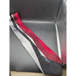 May include: A patterned necktie featuring a design of alternating colors. The tie has sections of black, gray, and red. The red section has a textured pattern. The tie is made of a smooth fabric.