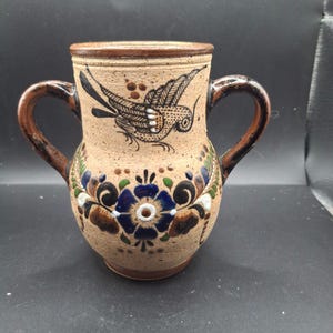 Puede incluir: Jarra de cerámica hecha a mano con dos asas. La jarra tiene una base beige con un borde y asas marrones. Está decorada con un diseño de pájaro negro y motivos florales en azul, verde y marrón. La jarra mide aproximadamente 15 cm de altura.
