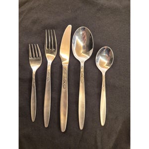 Puede incluir: Un juego de cinco cubiertos plateados, que incluye tenedores, un cuchillo y cucharas. Cada pieza tiene un diseño de hoja decorativa en el mango. Los cubiertos están dispuestos sobre un fondo oscuro.