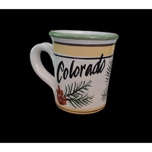 May include: White ceramic mug with a handle, featuring the word "Colorado" in black script. The mug is decorated with green pine needles and a brown pinecone. The rim has a green border, and the body has yellow and brown stripes.