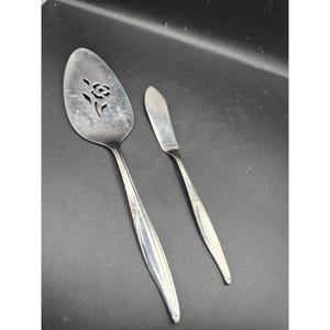 Puede incluir: Una cuchara de servir plateada con un diseño de corte de rosa y un cuchillo de mantequilla. Ambos utensilios tienen mangos largos y delgados con detalles decorativos. El juego es probablemente vintage.