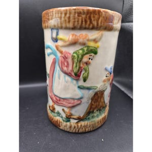 Puede incluir: Una taza de cerámica con borde y base marrón claro, con un diseño en relieve de figuras caprichosas en tonos rosa, verde y azul. La taza tiene un cuerpo color crema y una forma cilíndrica.