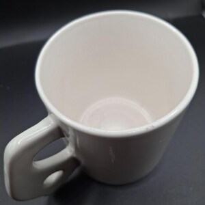 May include: A white ceramic coffee mug with a rounded body and a large, curved handle. The mug has a smooth, glossy finish and a simple, minimalist design. The interior of the mug is also white.