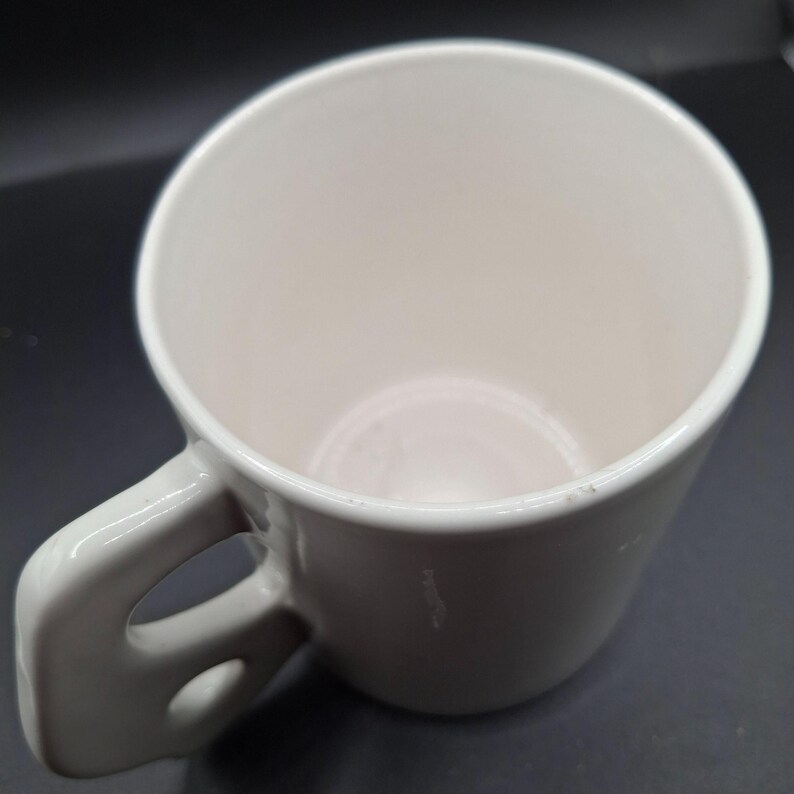 May include: A white ceramic coffee mug with a rounded body and a large, curved handle. The mug has a smooth, glossy finish and a simple, minimalist design. The interior of the mug is also white.