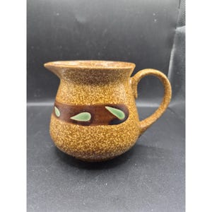 May include: A ceramic jug with a speckled tan exterior and a brown band featuring green leaf-shaped designs. The jug has a handle and a spout for pouring.
