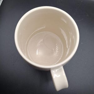 K&ouml;nnte beinhalten: Eine cremefarbene Keramiktasse mit gebogenem Henkel. Die Tasse hat eine glatte, gl&auml;nzende Oberfl&auml;che und eine zylindrische Form. Das Innere der Tasse ist sichtbar, und eine Haarstr&auml;hne befindet sich im Inneren. Die Tasse steht auf einer dunklen Oberfl&auml;che.
