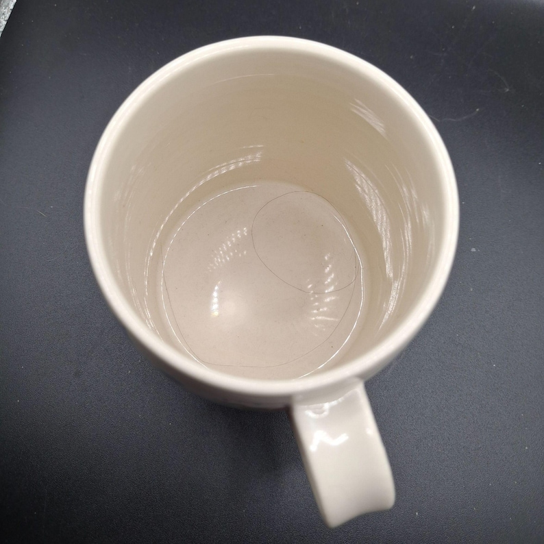 K&ouml;nnte beinhalten: Eine cremefarbene Keramiktasse mit gebogenem Henkel. Die Tasse hat eine glatte, gl&auml;nzende Oberfl&auml;che und eine zylindrische Form. Das Innere der Tasse ist sichtbar, und eine Haarstr&auml;hne befindet sich im Inneren. Die Tasse steht auf einer dunklen Oberfl&auml;che.