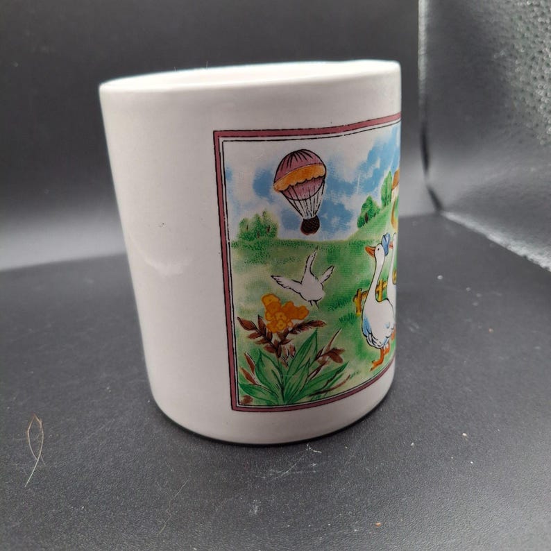 May include: White ceramic mug with a colorful illustration of a hot air balloon, geese, and flowers. The mug has a slightly tapered shape and a simple design, perfect for enjoying your favorite beverage.