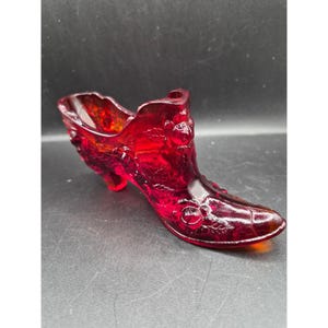 May include: A ruby red glass shoe figurine with floral detailing. The decorative shoe has a high heel and is made of transparent glass. The shoe is a decorative item, possibly a collectible or a small container.