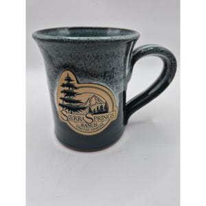 Op de afbeelding: Een keramische mok met een donkerblauwe en grijze gespikkelde glazuurlaag en een beige embleem aan de voorzijde. Het embleem toont een zwarte afbeelding van een dennenboom en bergen met de tekst SIERRA SPRINGS RANCH PINETOP, ARIZONA in het zwart.