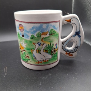 May include: White ceramic mug with a whimsical countryside scene featuring geese, a house, and a hot air balloon. The handle is shaped like a goose. The mug has a decorative border and is suitable for hot or cold beverages.