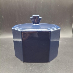 May include: A navy blue, octagonal ceramic container with a matching lid and a decorative handle. The container has a glossy finish and is likely used for storage or display. The design is simple and elegant.