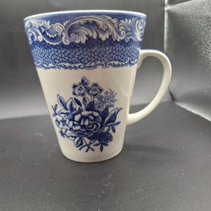 May include: White ceramic mug with a blue floral design and a decorative blue border. The mug has a curved handle and a flared shape. The floral design features detailed blue flowers and leaves. The mug is suitable for hot or cold beverages.