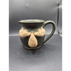 May include: Dark green ceramic pitcher with a handle and a spout. The pitcher features a face design with tan-colored clay accents. The face includes two cloud-like shapes and a teardrop shape.
