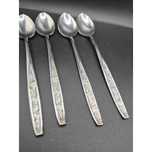May include: Four stainless steel spoons with a floral design on the handles. The spoons have a rounded bowl and a long, slender handle. The spoons are arranged in a row against a dark background.