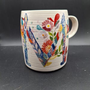 K&ouml;nnte beinhalten: Cremefarbene Keramiktasse mit einem gro&szlig;en, farbenfrohen floralen "M"-Design. Die Blumen sind in Rot-, Blau-, Gelb- und Gr&uuml;nt&ouml;nen gehalten. Die Tasse hat eine gerippte Textur und einen gebogenen Henkel.