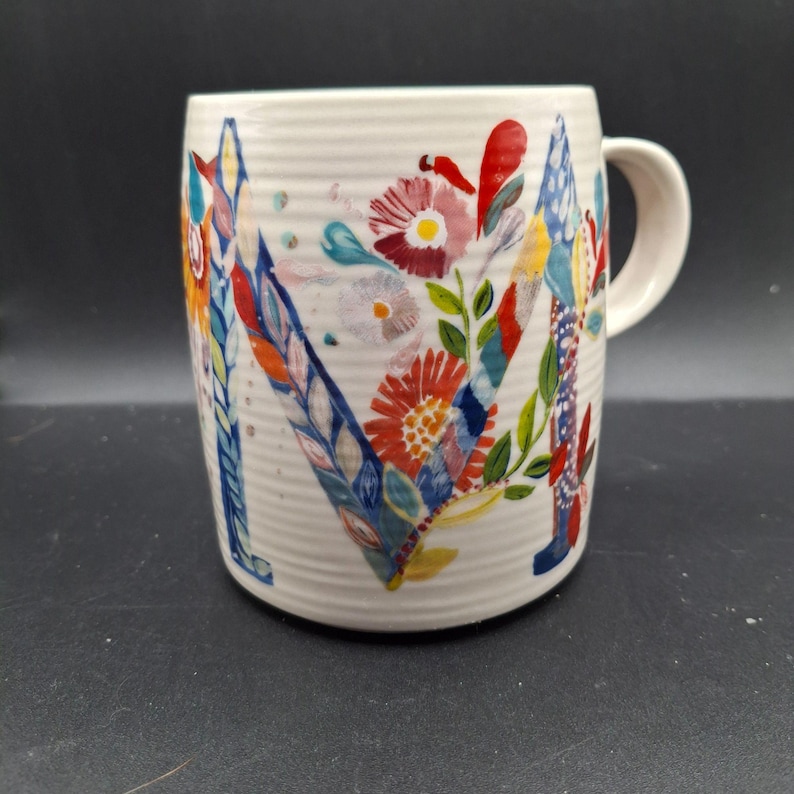K&ouml;nnte beinhalten: Cremefarbene Keramiktasse mit einem gro&szlig;en, farbenfrohen floralen "M"-Design. Die Blumen sind in Rot-, Blau-, Gelb- und Gr&uuml;nt&ouml;nen gehalten. Die Tasse hat eine gerippte Textur und einen gebogenen Henkel.