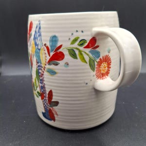 K&ouml;nnte beinhalten: Wei&szlig;e Keramiktasse mit floralem Design in Rot, Blau, Gelb und Gr&uuml;n. Die Tasse hat eine gerippte Textur und einen gebogenen Henkel. Das florale Design umfasst Bl&auml;tter und Blumen.