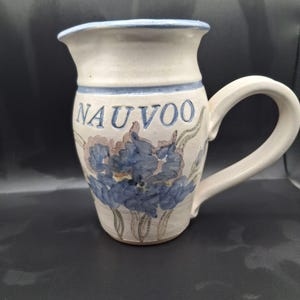 May include: Cream-colored ceramic pitcher with a large handle and a flared top. The pitcher features blue floral designs and the word "NAUVOO" in blue lettering. A blue band accents the top rim.