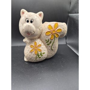 May include: A ceramic squirrel figurine with a planter opening. The squirrel is light beige with a smiling face, black eyes, and pink accents. Yellow flowers with green leaves decorate the body and tail. A whimsical decorative piece.