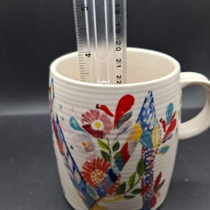 K&ouml;nnte beinhalten: Eine wei&szlig;e Keramiktasse mit einem bunten Blumendesign. Die Tasse hat einen Henkel und eine geriffelte Textur. Ein Lineal zeigt, dass die Tasse etwa 10 cm hoch ist.