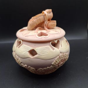 May include: A decorative trinket box in shades of pink and cream, featuring a pair of ballet slippers on the lid. The box has heart-shaped cutouts and swan-like designs, with a textured base.