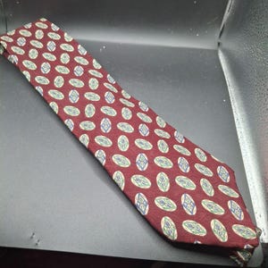 May include: A burgundy tie with a repeating pattern of oval shapes in green, blue, and white. The tie is made of a smooth fabric and is displayed on a dark surface.