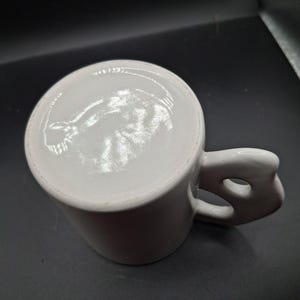 May include: White ceramic mug with a unique handle design. The handle features a distinctive cutout, adding a modern touch to the classic mug shape. The mug is clean and simple, perfect for coffee or tea.