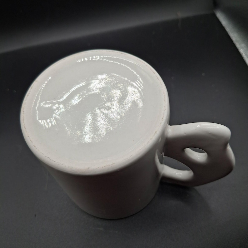 May include: White ceramic mug with a unique handle design. The handle features a distinctive cutout, adding a modern touch to the classic mug shape. The mug is clean and simple, perfect for coffee or tea.