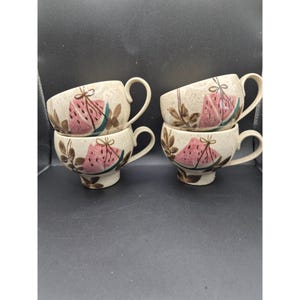 Könnte beinhalten: Vier Keramik-Kaffeetassen, die paarweise gestapelt sind. Jede Tasse hat einen beigefarbenen, gesprenkelten Hintergrund mit einem rosa Wassermelonen-Scheiben-Design, braunen Blättern und einem gebogenen Henkel. Die Tassen stehen auf einer dunklen Oberfläche.
