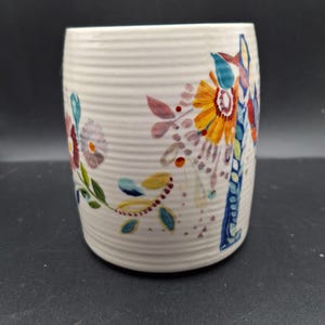 K&ouml;nnte beinhalten: Eine wei&szlig;e Keramiktasse mit gerippter Textur und farbenfrohen Blumendesigns. Die Blumen sind in Orange-, Gelb-, Blau- und Rott&ouml;nen bemalt, mit gr&uuml;nen Bl&auml;ttern und Akzenten. Die Tasse hat eine zylindrische Form.
