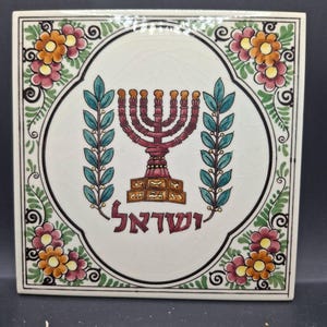 May include: Square ceramic tile featuring a central design of a maroon menorah with gold accents, surrounded by teal leaves and floral patterns in pink and orange. The Hebrew word "Israel" is written in maroon. The tile has a white background and a black border.